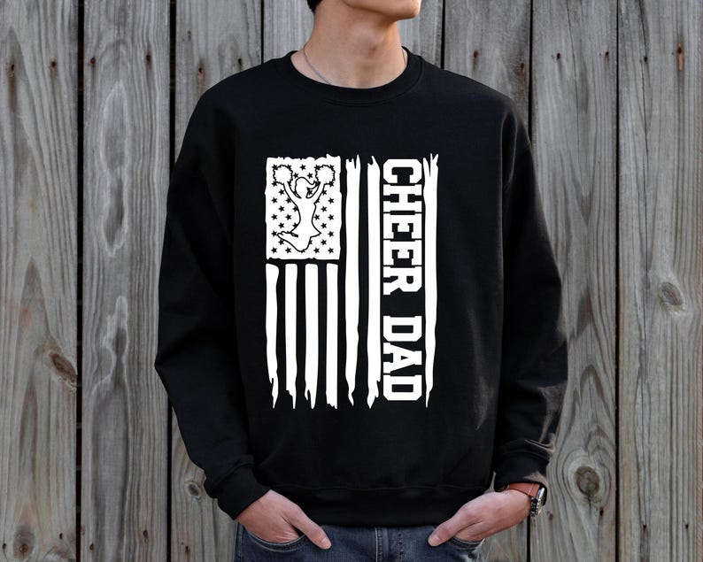 May include: Black sweatshirt with a white distressed American flag design. The flag features a cheerleader silhouette and the text "Cheer Dad".
