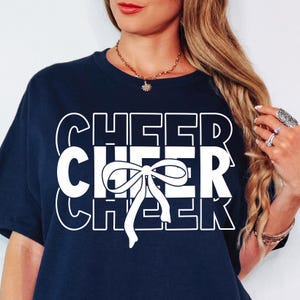 Pode incluir: Uma camiseta azul marinho com um design gráfico branco que diz "CHEER" três vezes com um laço branco no meio.