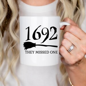 May include: White ceramic mug with black text and a black silhouette of a broom. The text reads "1692 They Missed One".