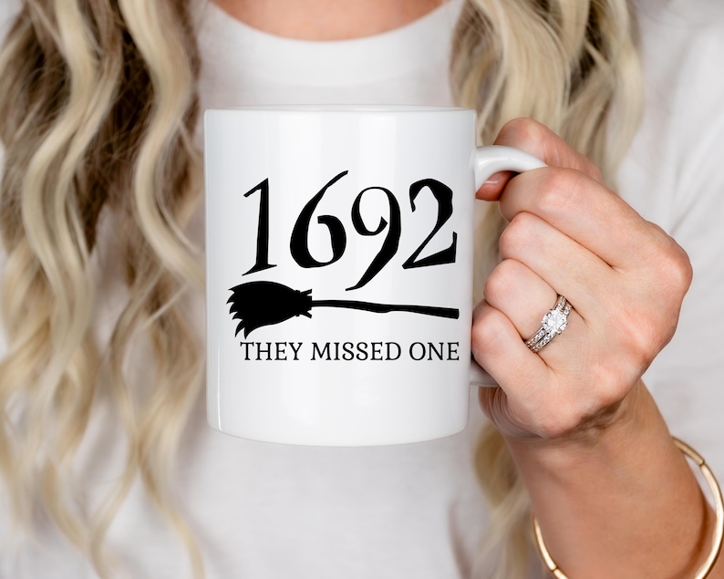 May include: White ceramic mug with black text and a black silhouette of a broom. The text reads "1692 They Missed One".