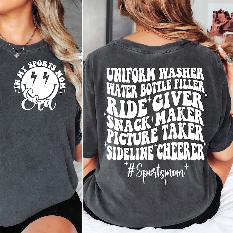 May include: A dark gray t-shirt with a white graphic on the back that says "Uniform Washer, Water Bottle Filler, Ride Giver, Snack Maker, Picture Taker, Sideline Cheerer #Sportsmom". The front of the shirt has a white graphic of a smiley face with lightning bolts and the text "I'm My Sports Mom Era".
