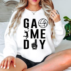 May include: White sweatshirt with the words "GAME DAY" in large black letters. The design includes a volleyball with a heart, a volleyball player, and a foam finger. A casual, sporty top.