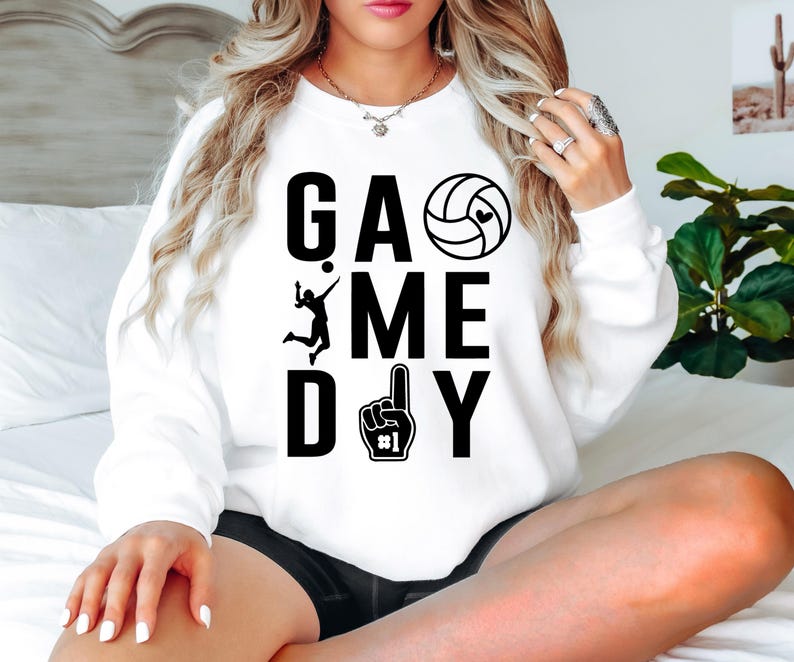 May include: White sweatshirt with the words "GAME DAY" in large black letters. The design includes a volleyball with a heart, a volleyball player, and a foam finger. A casual, sporty top.