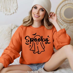 Op de afbeelding: Een vrouw draagt een oranje trui met een zwarte geest en de tekst "Spooky" erop.