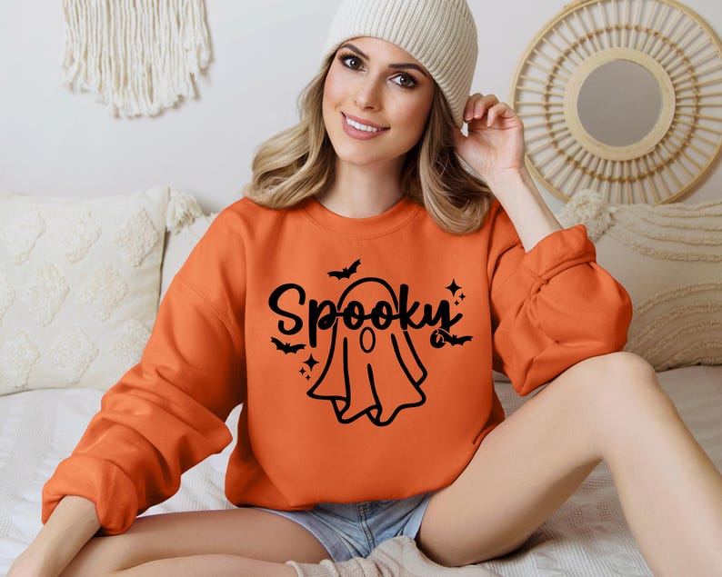 Op de afbeelding: Een vrouw draagt een oranje trui met een zwarte geest en de tekst "Spooky" erop.