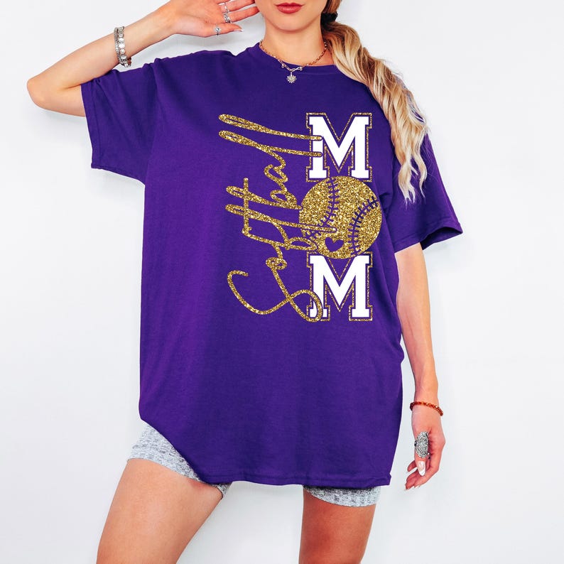 May include: A purple t-shirt with a gold glitter design. The design features the word "MOM" with a baseball in the center. The baseball is gold glitter, and the letters "M" are white.