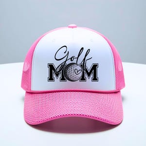 Może przedstawiać: Biało-r&oacute;żowa czapka trucker z czarno-białym nadrukiem z napisem "Golf Mom" z wzorem piłki golfowej i serca.