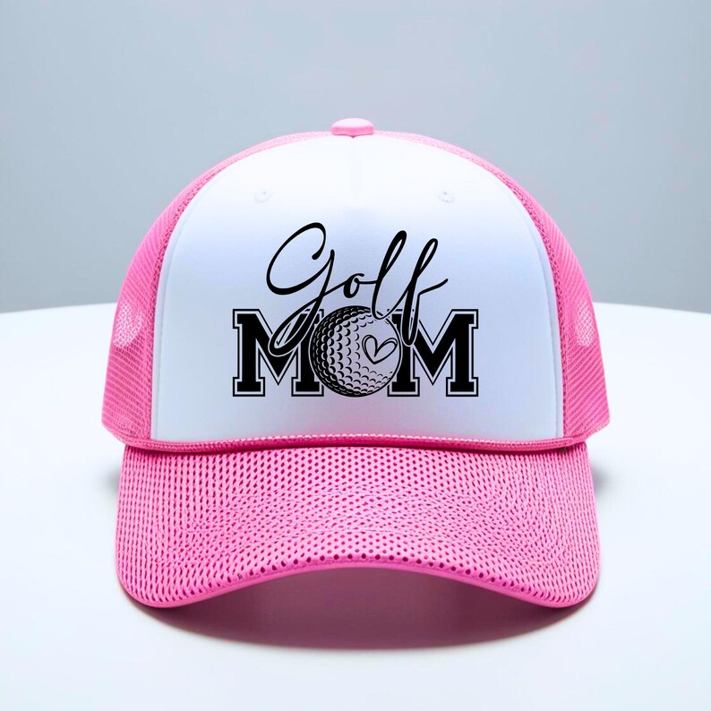 Może przedstawiać: Biało-r&oacute;żowa czapka trucker z czarno-białym nadrukiem z napisem "Golf Mom" z wzorem piłki golfowej i serca.