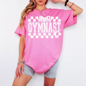 May include: Pink t-shirt with a white checkered pattern, the word "GYMNAST" in white block letters, and a white silhouette of a gymnast on the uneven bars.
