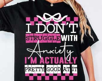 Puede incluir: Camiseta negra con un diseño a cuadros rosa y blanco. El texto "No lucho con la ansiedad, en realidad soy bastante buena en eso" está impreso en la camiseta.