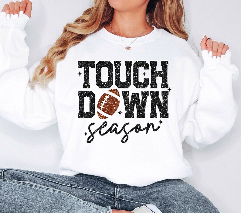 May include: White sweatshirt with black glitter text that reads "Touch Down Season" with a brown football graphic in the center.