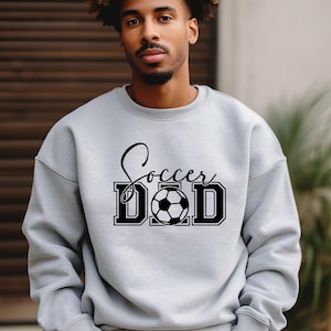 Peut inclure: Un sweat-shirt gris avec le texte "Soccer Dad" en lettres noires et un graphique de ballon de football noir et blanc.