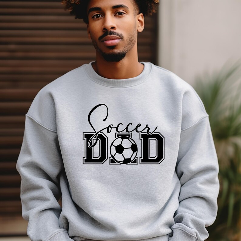 Peut inclure: Un sweat-shirt gris avec le texte "Soccer Dad" en lettres noires et un graphique de ballon de football noir et blanc.