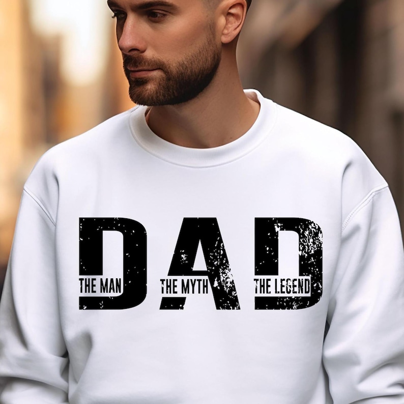 K&ouml;nnte beinhalten: Ein wei&szlig;es Sweatshirt mit Rundhalsausschnitt und dem schwarzen Schriftzug DAD in einer abgenutzten, verwitterten Schriftart. Darunter sind die W&ouml;rter THE MAN, THE MYTH und THE LEGEND in einer kleineren, klaren schwarzen Schrift zentriert abgebildet.
