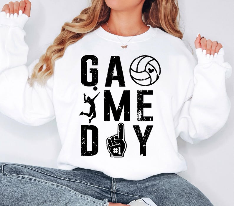 May include: White sweatshirt featuring the text "GAME DAY" in large, distressed black letters. A volleyball graphic and a foam finger with the number one are also present. The sweatshirt is a casual, sporty style.