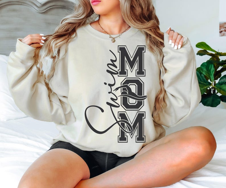 May include: A cream colored sweatshirt with the text "Swim Mom" in black lettering. The text is in a varsity style font with a wave design.