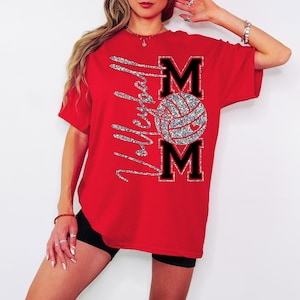 May include: A red t-shirt with the words "Volleyball Mom" in silver glitter script. The design includes a volleyball and the word "MOM" in black block letters with a silver glitter outline. The shirt is short-sleeved.
