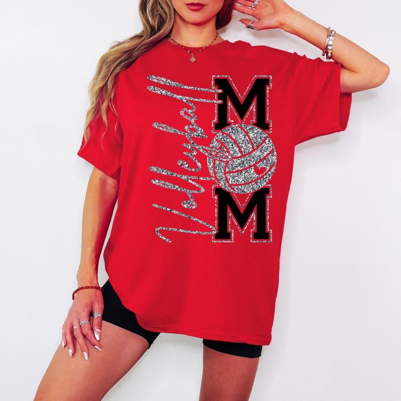 May include: A red t-shirt with the words "Volleyball Mom" in silver glitter script. The design includes a volleyball and the word "MOM" in black block letters with a silver glitter outline. The shirt is short-sleeved.