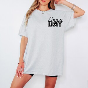May include: A light gray t-shirt with the words "Game Day" in black script and block letters, with a football graphic. The shirt is a casual, oversized style.