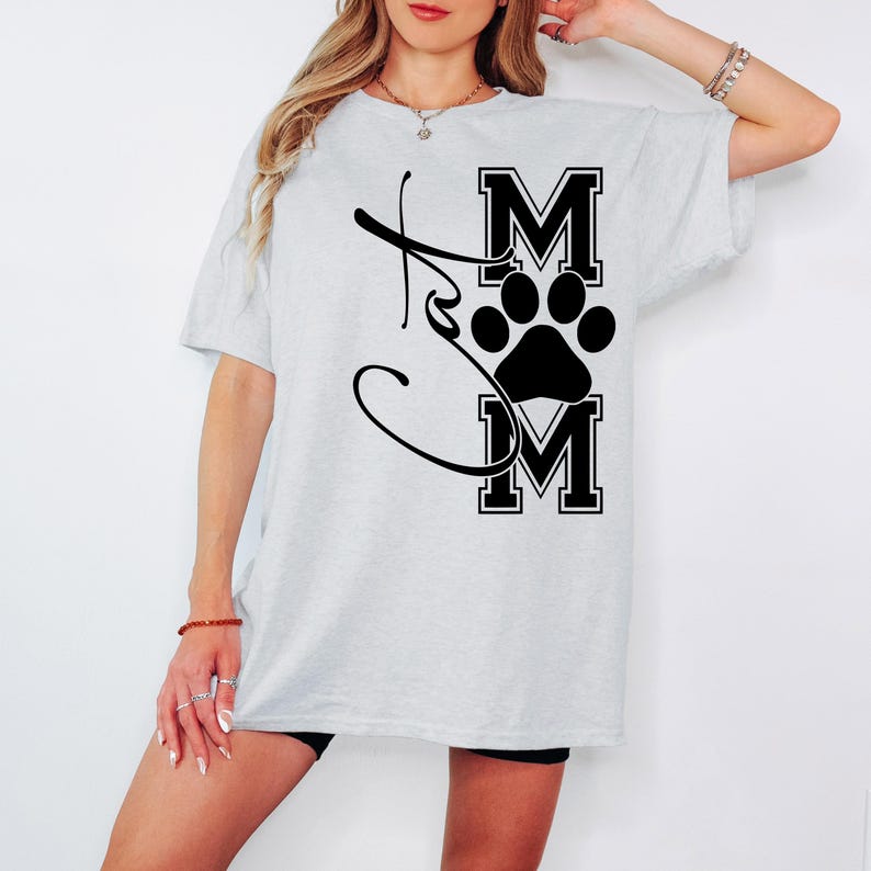 May include: Light gray t-shirt with a black graphic that says "Mom" with a paw print in the center. The word "Mom" is in a bold, block letter font. The shirt is a casual, oversized style.