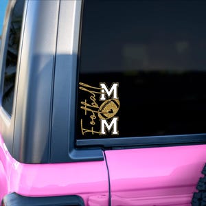 May include: A gold glitter vinyl decal on a vehicle window. The decal reads "Football Mom" with a football graphic between the words. The vehicle is pink and parked on a sandy beach with palm trees in the background.