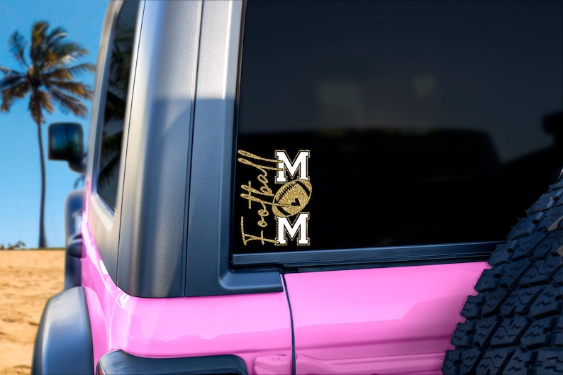 May include: A gold glitter vinyl decal on a vehicle window. The decal reads "Football Mom" with a football graphic between the words. The vehicle is pink and parked on a sandy beach with palm trees in the background.