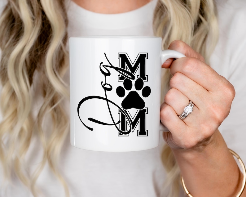 Puede incluir: Taza de caf&eacute; de cer&aacute;mica blanca con una huella de pata negra y el texto "Mom" en una fuente negra en bloque.