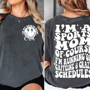 Pode incluir: Uma camiseta cinza escura com um gr&aacute;fico branco na frente que diz "In My Sports Mom Era" com um rosto sorridente e raios. A parte de tr&aacute;s da camiseta tem um gr&aacute;fico branco que diz "I'm a Sports Mom of Course I'm Running on Caffeine & Crazy Schedules".