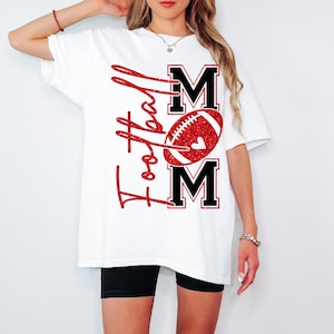 May include: White t-shirt with a red glitter design that says "Football Mom". The design includes a football and the word "MOM" in black block letters. The shirt is a casual, oversized fit.