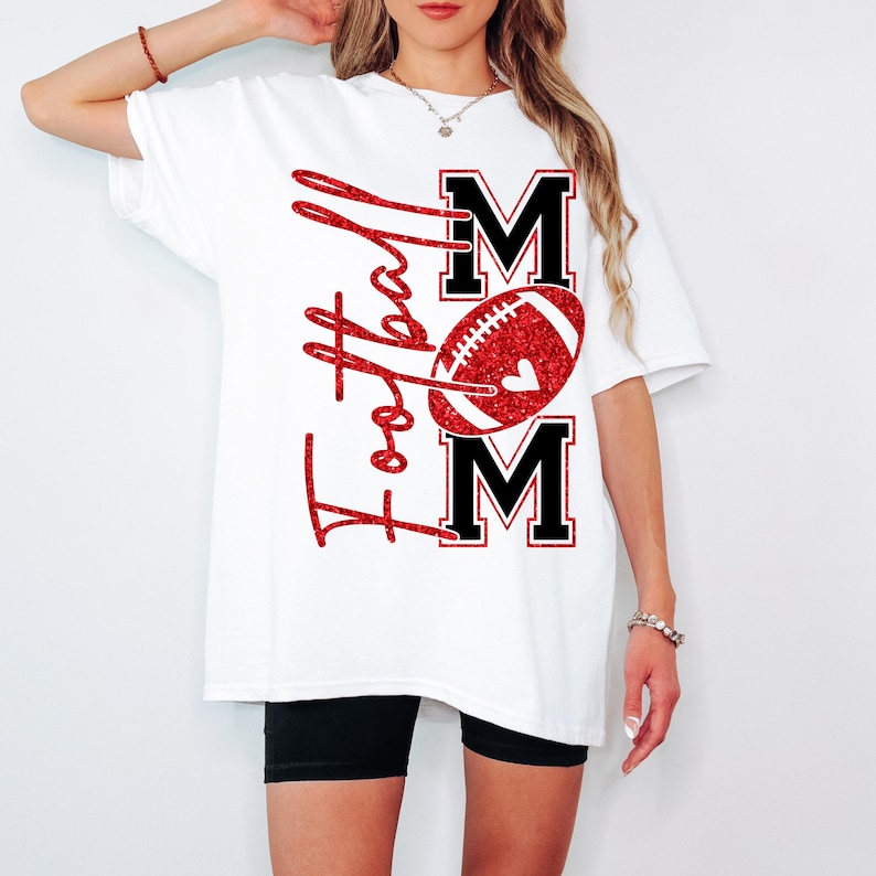 May include: White t-shirt with a red glitter design that says "Football Mom". The design includes a football and the word "MOM" in black block letters. The shirt is a casual, oversized fit.