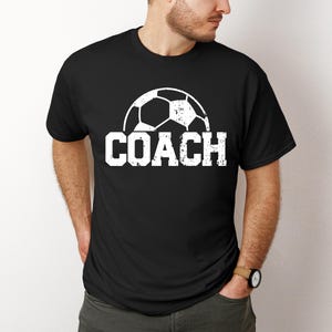 May include: Black t-shirt with a white soccer ball graphic and the word "COACH" in white distressed lettering.