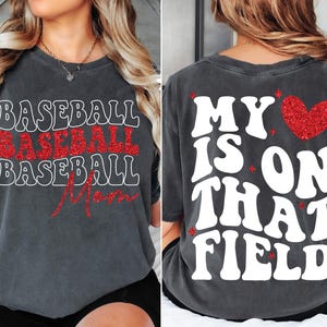 Pu&ograve; includere: Una maglietta grigia con il testo "Baseball Mom" in glitter rosso sul davanti e "My heart is on that field" in bianco sul retro con un cuore rosso glitterato.