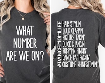 May include: A dark grey t-shirt with white text that reads "WHAT NUMBER ARE WE ON?" on the front and "#DANCEMOMLIFE HAIR STYLIN LOUD CLAPPIN PICTURE TAKIN QUICK CHANGIN BOBBYPIN FINDIN DANCE BAG PACKIN COSTUME RHINESTONIN" on the back.