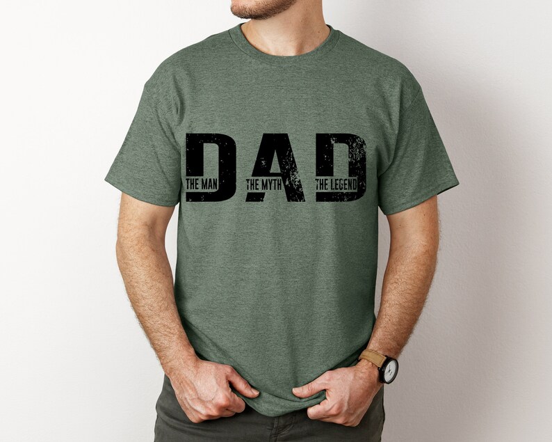 K&ouml;nnte beinhalten: Ein kurz&auml;rmeliges T-Shirt in meliertem Olivgr&uuml;n mit dem schwarzen, abgenutzten Schriftzug DAD auf der Brust. Innerhalb der Buchstaben des Wortes DAD sind die Phrasen THE MAN, THE MYTH und THE LEGEND in einer kleineren schwarzen Schrift gedruckt.
