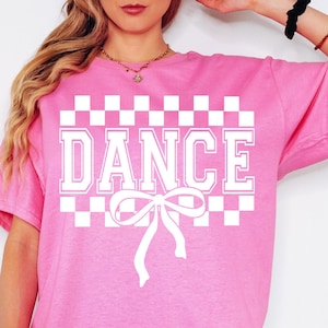 May include: Pink t-shirt with a white checkered pattern and the word "DANCE" in white block letters. A white bow is centered below the word.