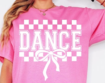 May include: Pink t-shirt with a white checkered pattern and the word "DANCE" in white block letters. A white bow is centered below the word.