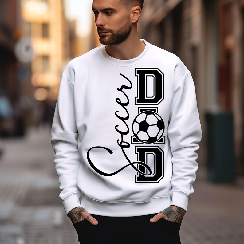 Peut inclure: Sweat-shirt blanc avec un texte noir qui dit "Soccer Dad" avec un graphique de ballon de football.