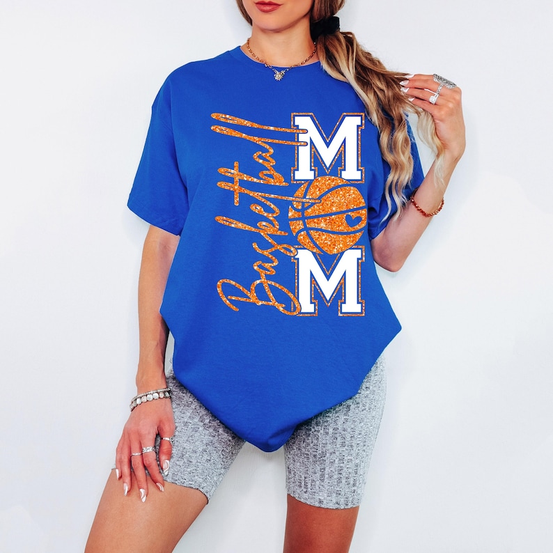 May include: Royal blue t-shirt with the words "Basketball Mom" in glittery orange script. The design includes a basketball and the letters "M" above and below the ball in white. The shirt is a casual style.