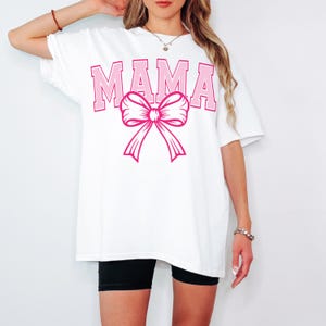 May include: White oversized t-shirt with the word "MAMA" in pink letters and a pink bow.