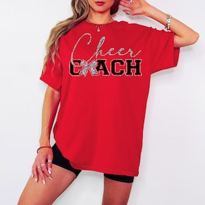 May include: A red t-shirt featuring the text "Cheer Coach". The word "Cheer" is in silver glitter script, and "COACH" is in black block letters. The shirt has a round neck and short sleeves. Cheerleading theme.