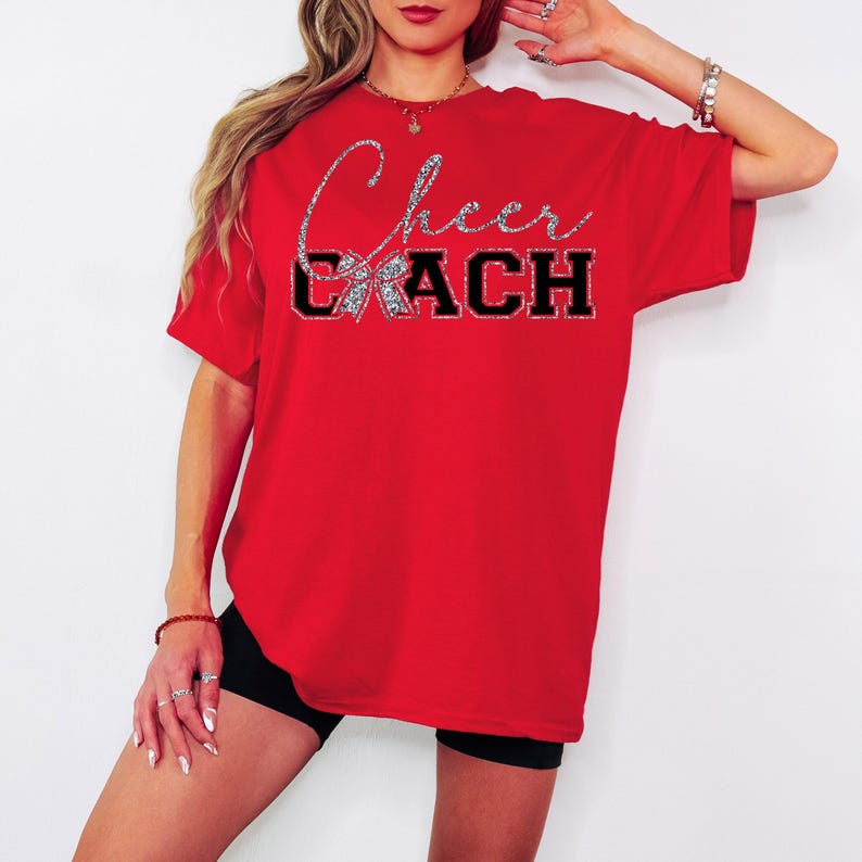 May include: A red t-shirt featuring the text "Cheer Coach". The word "Cheer" is in silver glitter script, and "COACH" is in black block letters. The shirt has a round neck and short sleeves. Cheerleading theme.