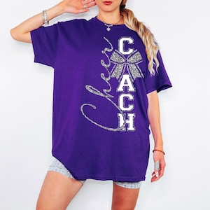 May include: A purple t-shirt featuring the word "Coach" in white block letters and a silver glitter bow graphic. The word "Cheer" is written in a silver glitter script. The shirt has a relaxed, oversized fit.
