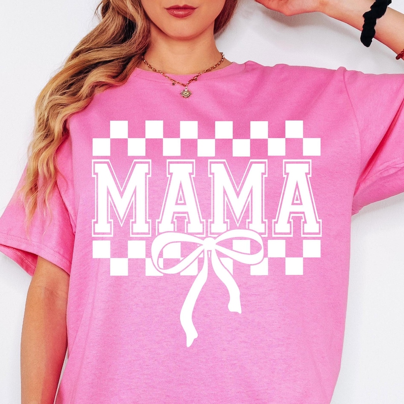 May include: Pink t-shirt with a white checkered pattern and the word "MAMA" in white block letters. A white bow is centered below the word.