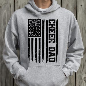 May include: A grey hooded sweatshirt with a black distressed American flag design. The flag features a cheerleader silhouette and the text "CHEER DAD" on the right side.
