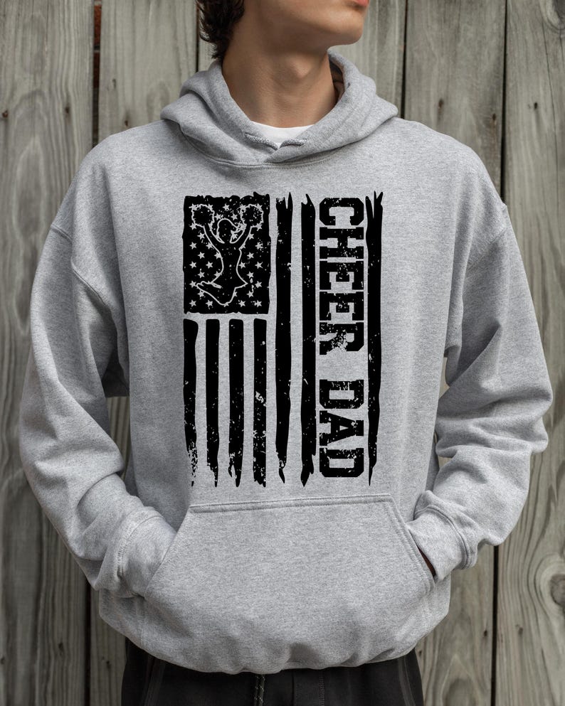 May include: A grey hooded sweatshirt with a black distressed American flag design. The flag features a cheerleader silhouette and the text "CHEER DAD" on the right side.