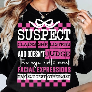 Op de afbeelding: Zwart sweatshirt met een roze en wit geruit patroon en de tekst "SUSPECT CLAIMS SHE LISTENS AND DOESN'T JUDGE the eye rolls and FACIAL EXPRESSIONS MAY SUGGEST OTHERWISE".
