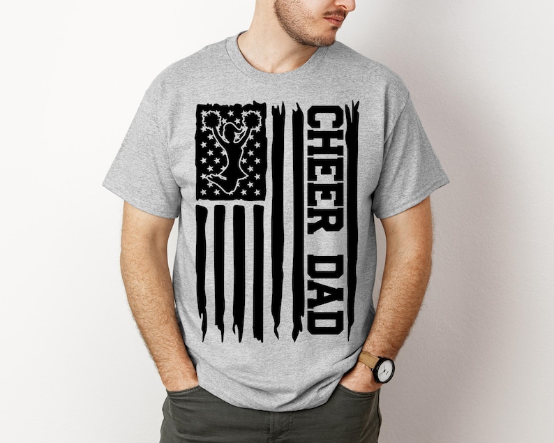 May include: A grey t-shirt with a black and white American flag design. The flag features a silhouette of a cheerleader with pom-poms and the text "Cheer Dad".