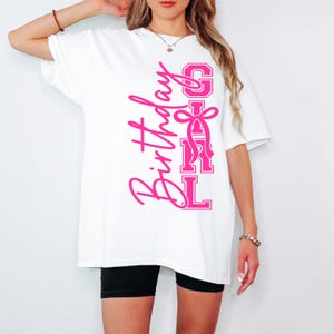 May include: White t-shirt featuring the text "Birthday Girl" in pink script and block letters. The design includes a pink bow. The shirt is worn with black shorts. The t-shirt is a casual style.