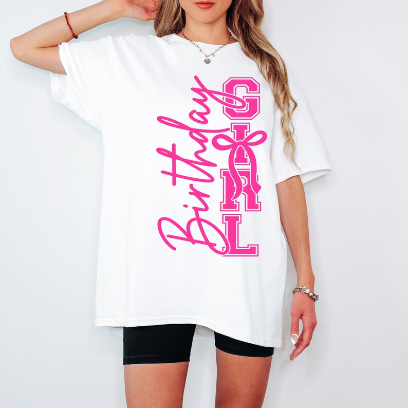 May include: White t-shirt featuring the text "Birthday Girl" in pink script and block letters. The design includes a pink bow. The shirt is worn with black shorts. The t-shirt is a casual style.