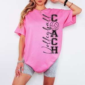 K&ouml;nnte beinhalten: Pinkes T-Shirt mit einem schwarzen Grafikdesign, auf dem "Volleyball Coach" mit einem Volleyball und einem Herz steht.